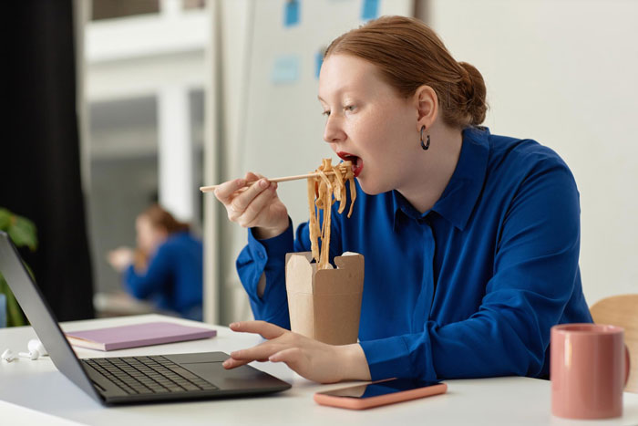 Woman eating takeout noodles while working on laptop, illustrating workers adding labels to protect their food from lunch thieves.