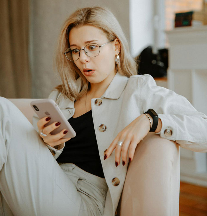 Young woman with glasses looking shocked at phone, illustrating awareness of romance scam risk in coworker conversation. Young woman with glasses looking shocked at phone, illustrating awareness of romance scam risk in coworker conversation.