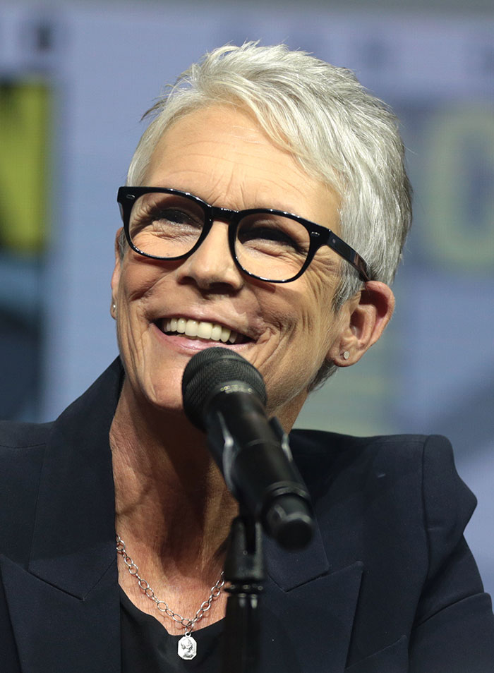 Smiling woman with short gray hair and glasses speaking into a microphone about celebrity rumors at an event.