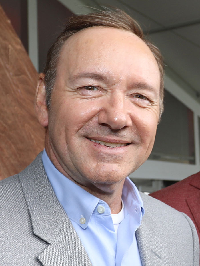 Middle-aged man smiling indoors, wearing light blue shirt and gray blazer, illustrating wild celebrity rumors concept.