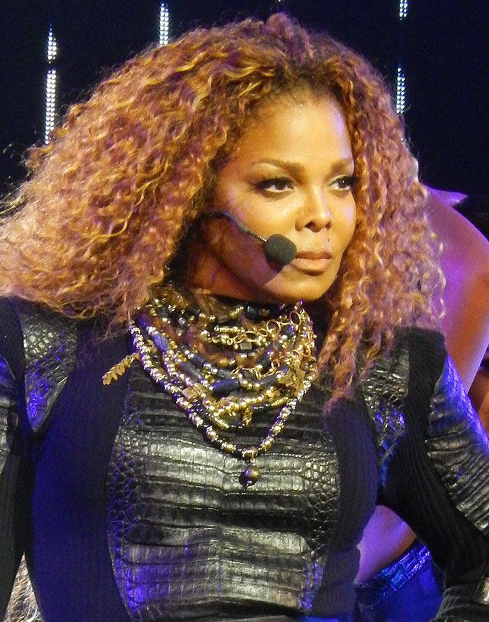 Performer with curly hair wearing a microphone headset and layered necklaces during a live celebrity event on stage.