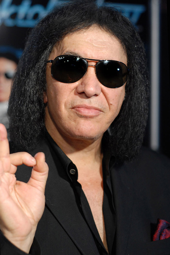 Man with black sunglasses and long curly hair making an OK hand gesture amid celebrity rumors and gossip background.