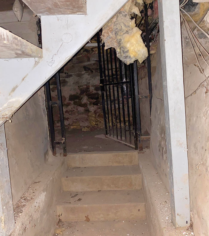Narrow dirty basement stairs leading to a hidden old room with a rusty metal gate and exposed insulation above.