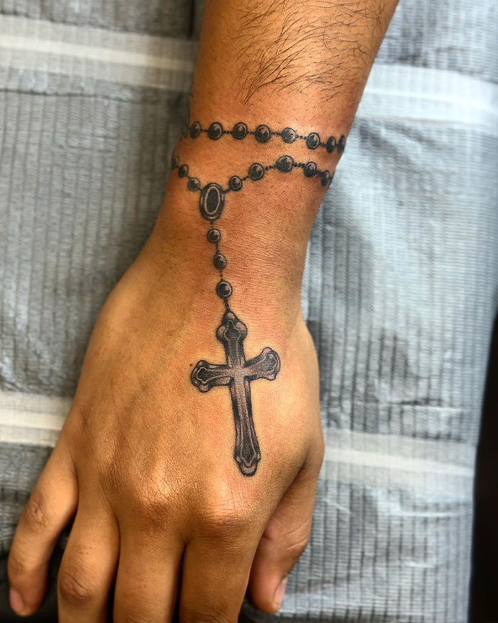 Close-up of a cross tattoo on a man's hand designed to resemble a rosary with detailed beads and shading.