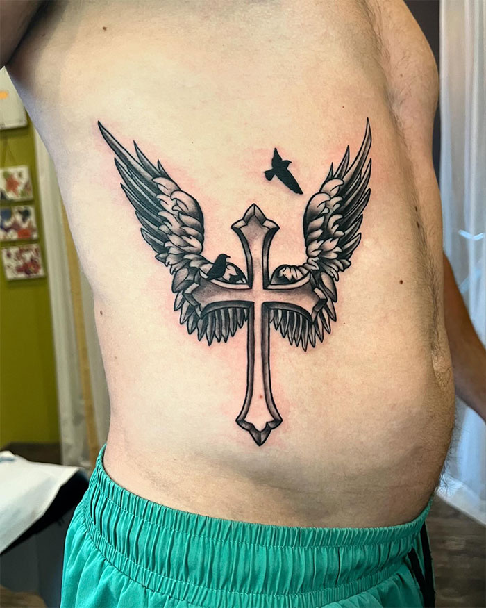 Cross tattoo with wings and birds inked on a man's side chest in black and gray shading style.