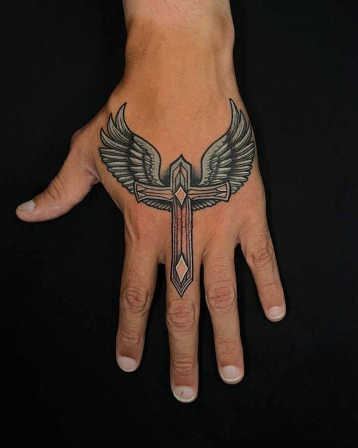 Hand tattoo of a detailed cross with wings, showcasing a popular cross tattoo design for men on a black background.