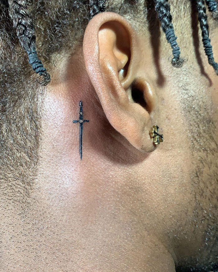 Small black cross tattoo behind the ear on a man with braided hair, showcasing cross tattoos for men.