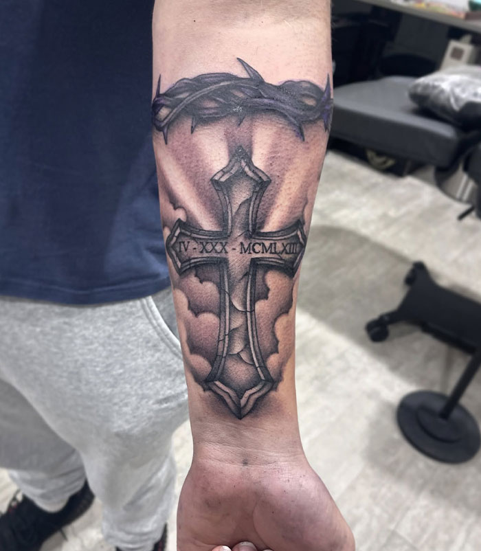 Forearm cross tattoo with Roman numerals and a crown of thorns, showcasing detailed shading in a men's tattoo design.