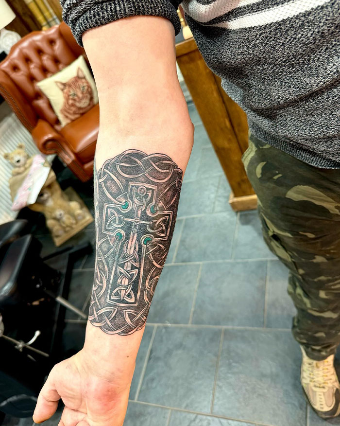 Intricate Celtic cross tattoo on a man's forearm showcasing detailed knotwork in a tattoo studio setting.