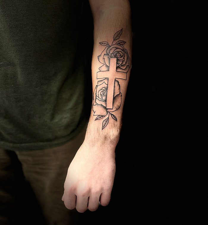 Forearm cross tattoo with roses and leaves in black ink on a man's arm showcasing cross tattoos for men.