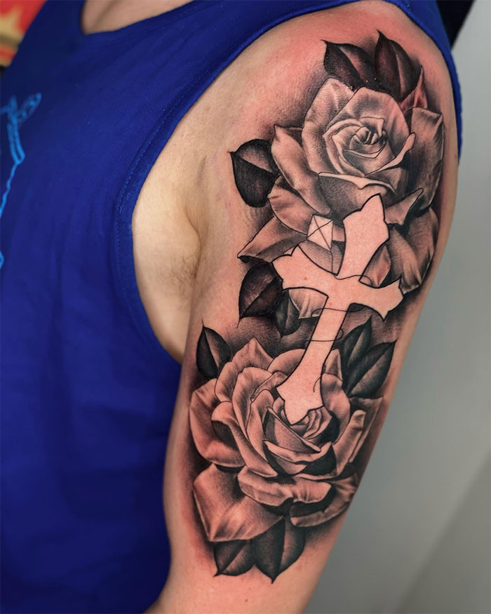 Black and grey cross tattoo surrounded by detailed roses on a man's upper arm, showcasing popular cross tattoo design for men.