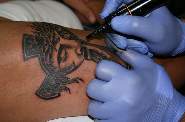 Close-up of a man getting detailed cross tattoo inked on his arm by a tattoo artist wearing blue gloves.