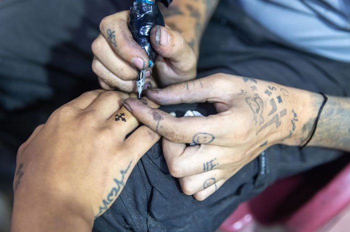Close-up of a tattoo artist creating a small cross tattoo on a man's finger in a detailed ink session.