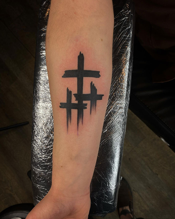 Black ink cross tattoos on a man's forearm with a bold and dripping paintbrush style design.