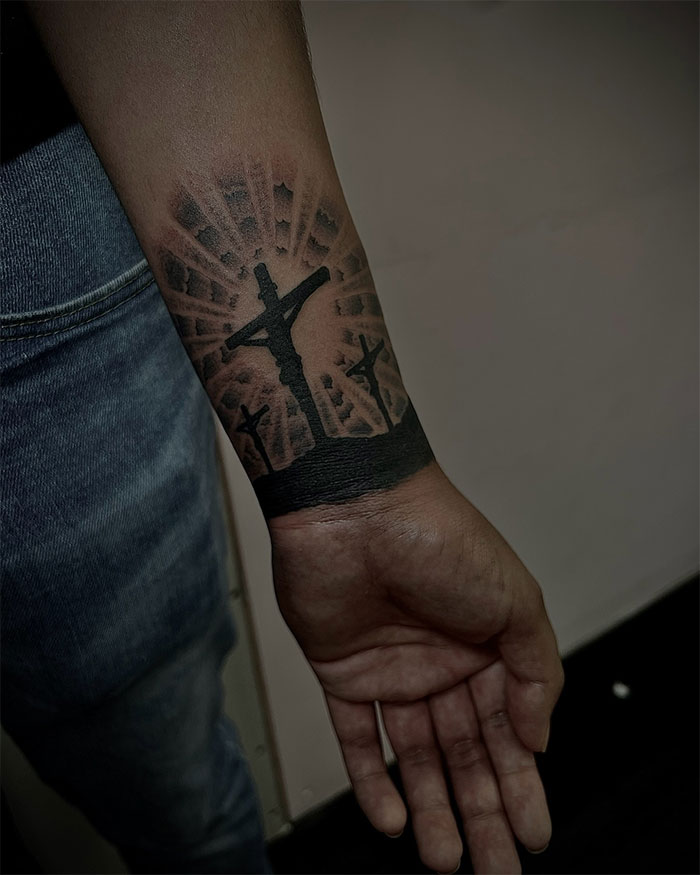 Black cross tattoo on man's forearm featuring three crosses with rays of light in the background, visual guide to cross tattoos for men.