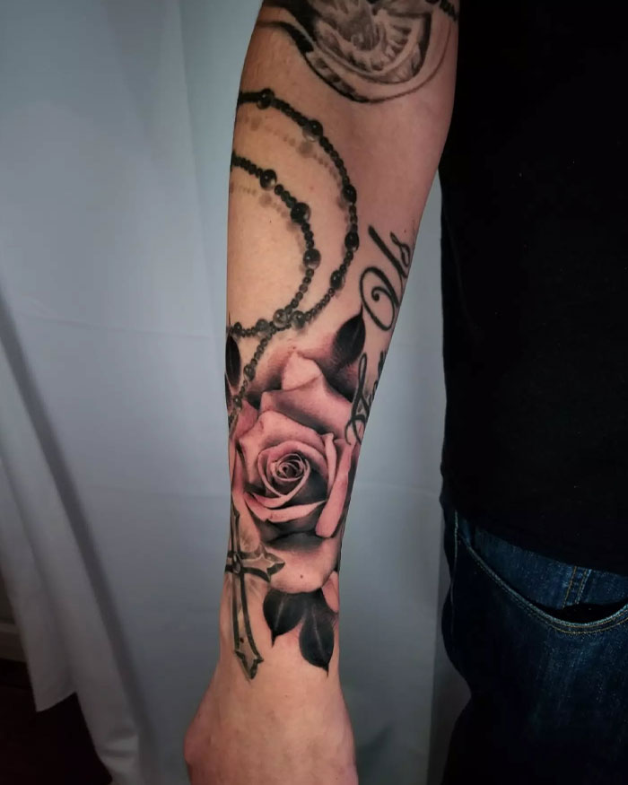 Forearm tattoo featuring a detailed cross, black rosary beads, and realistic roses in a men's cross tattoo design.