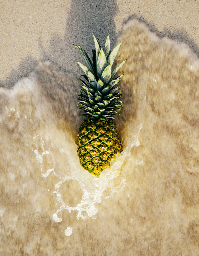 Pineapple on sandy beach with ocean waves washing over, symbolizing cruise passenger furious over pineapple shirt meaning.