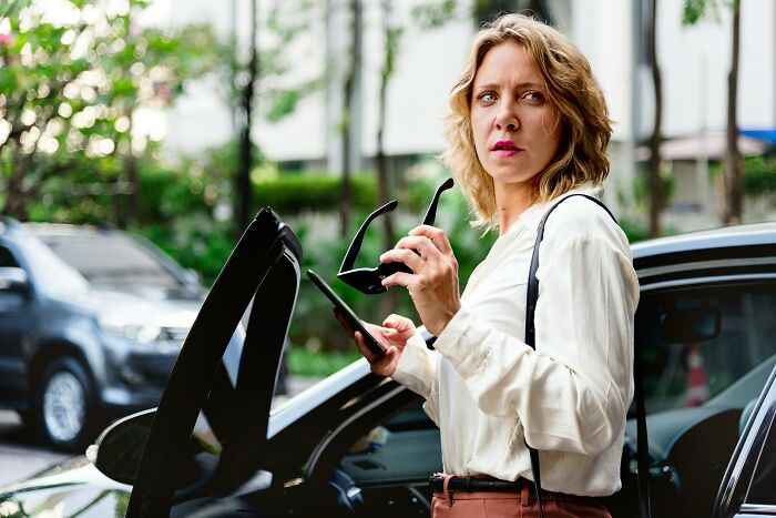 Woman looking upset holding sunglasses and phone by car door, illustrating unhinged ways people got back at cheating exes.