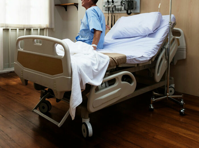 Patient in a hospital bed wearing a gown, sitting up and looking out the window in a quiet medical room.