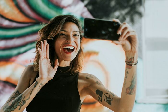 Young woman with tattoos smiling and waving while taking a selfie, illustrating behaviors that may indicate insecurity.
