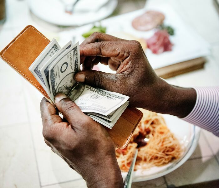 Hands holding a wallet counting cash bills over a table with a plate of spaghetti, illustrating poor people habits.
