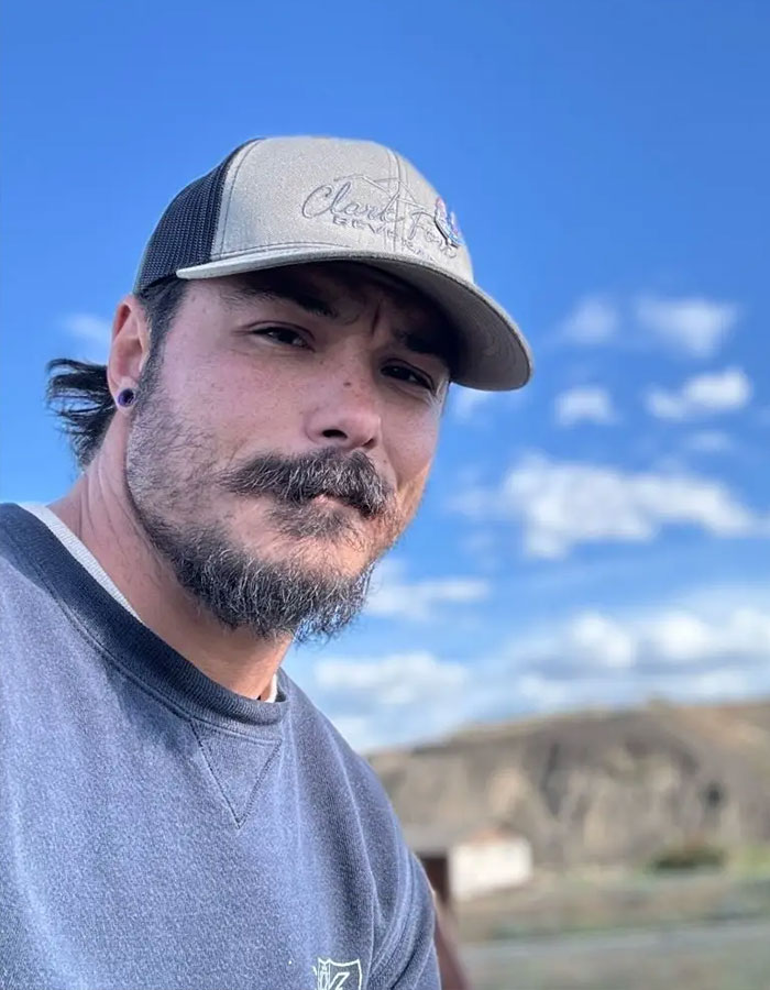 Man with beard wearing a cap outdoors under blue sky, featured in police search related to suffocated daughters case.