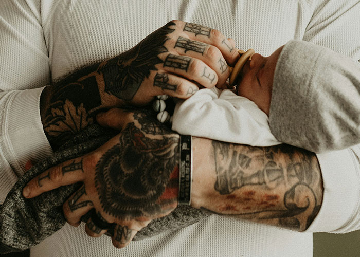 A tattooed parent tenderly holding a newborn baby, symbolizing emotional experiences of discovering kids aren’t biologically theirs.