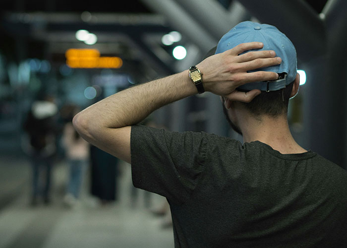 Man wearing a blue cap and black watch seen from behind, standing in a dimly lit urban environment reflecting broken family news.