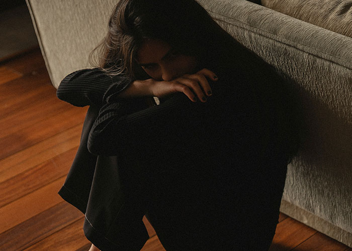 Woman sitting on wooden floor by sofa, appearing upset while reflecting on discovering kids weren’t biologically theirs.