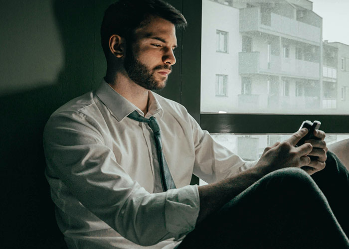 Man in a white shirt sitting by a window, looking distressed while holding a phone, reflecting on broken trust with kids.