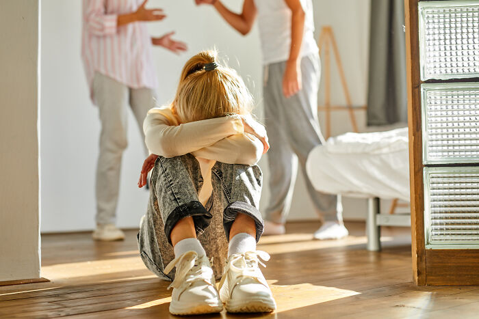 Child sitting on floor with head buried in arms while two adults argue in background, showing signs of difficult childhood.