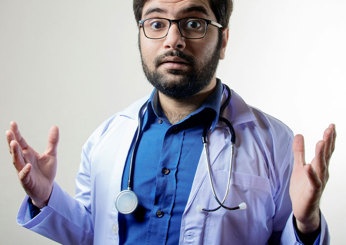 Male doctor with glasses and stethoscope shrugging in delivery room, illustrating stories about paternity and childbirth surprises.