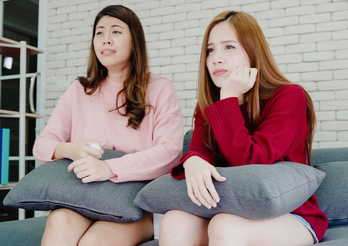Two women sitting on a couch holding pillows, appearing emotional and discussing delivery room stories about unclear fatherhood.