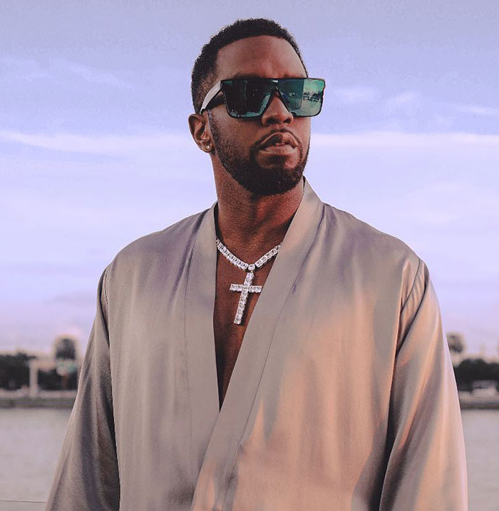 Diddy wearing sunglasses and a cross necklace, captured outdoors with a soft sunset background.