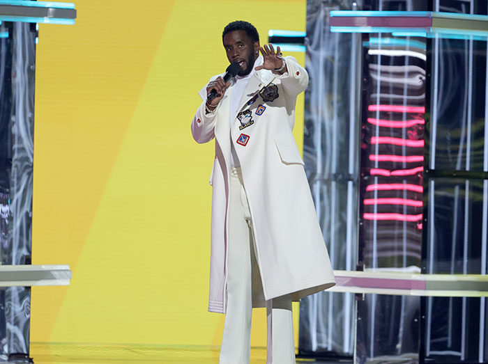 Male speaker wearing a white coat on stage, holding a microphone, in a vibrant setting related to Diddy court sketch artist coverage. Male speaker wearing a white coat on stage, holding a microphone, in a vibrant setting related to Diddy court sketch artist coverage.
