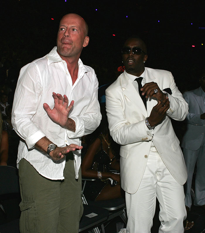Two men at an event, one in a white shirt and cargo pants, the other in a white suit and sunglasses, related to Diddy's trial.