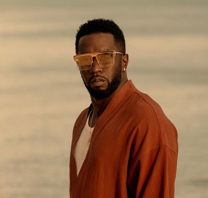 Man wearing amber sunglasses and rust-colored jacket posing outdoors with soft-focus background related to Diddy's trial witness.