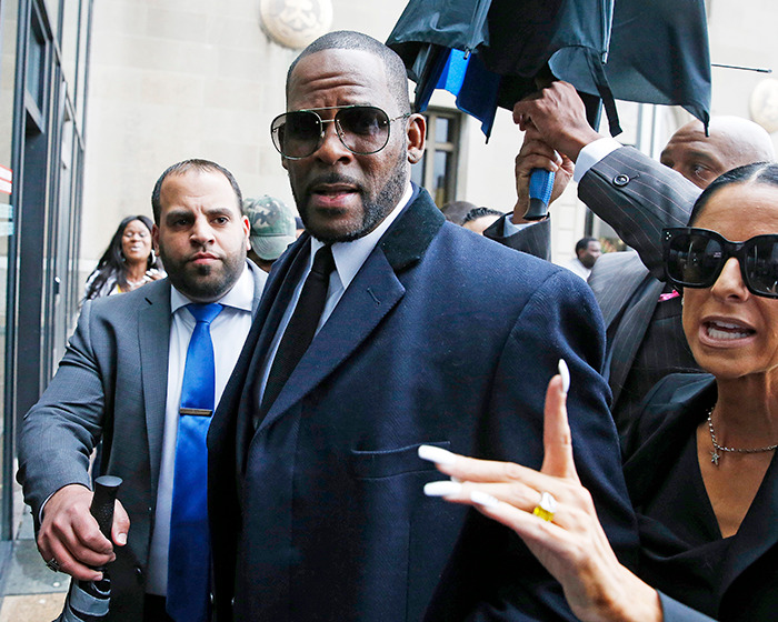 Rapper R Kelly wearing sunglasses and dark coat, surrounded by people outside a courthouse in a crowded setting. Rapper R Kelly wearing sunglasses and dark coat, surrounded by people outside a courthouse in a crowded setting.