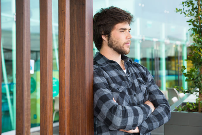 Young man in checkered shirt leaning against wooden wall, reflecting on friendship and job referral after divorce.