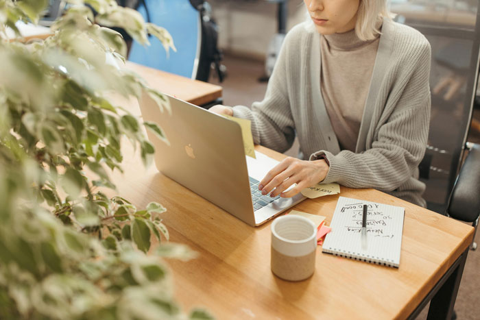 Woman working on a laptop at a desk, focused on typing, with notebook and coffee cup nearby in a cozy workspace.