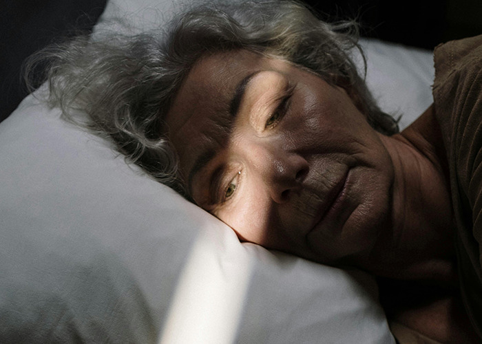 Elderly woman lying on a pillow with a shadow across her face, reflecting a medical case involving skin.