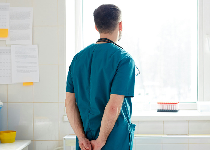 Medical professional in scrubs looking out window in a clinical setting, reflecting on bizarre medical cases witnessed.