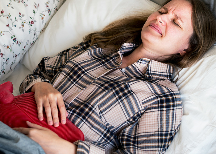 Woman in plaid shirt lying in bed holding hot water bottle on stomach showing signs of medical conditions that surprised doctors