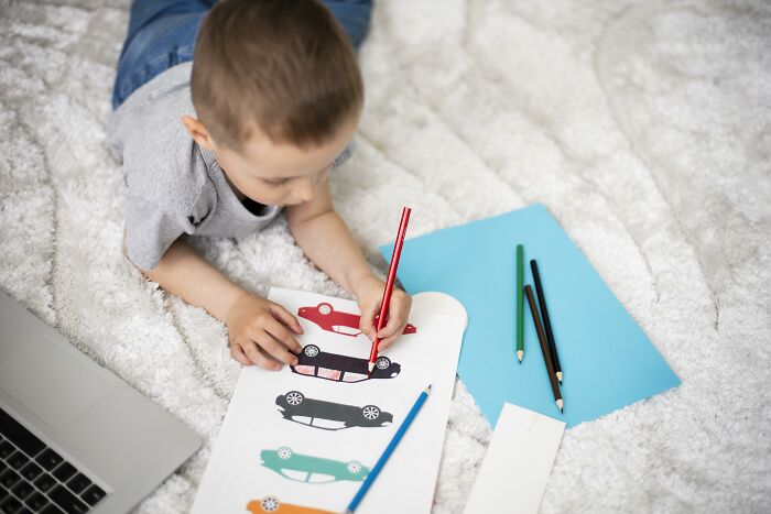 Young boy exploring creative freedom by coloring car drawings with colored pencils in an online art class setting.