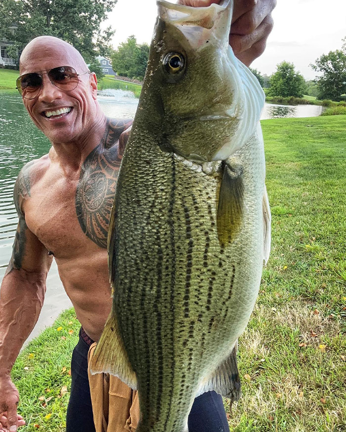 Dwayne Johnson smiling shirtless by a lake holding a large fish, showcasing his success and net worth growth.