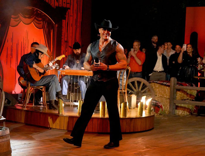 Dwayne Johnson performing in a cowboy hat and vest on stage with musicians, highlighting his rising net worth in 2025.