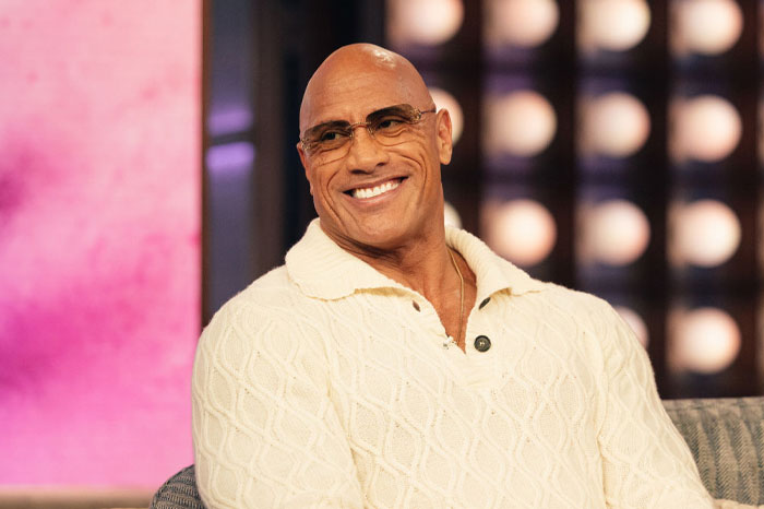Dwayne Johnson wearing glasses and a cream cable-knit sweater smiling during a television interview about net worth growth.