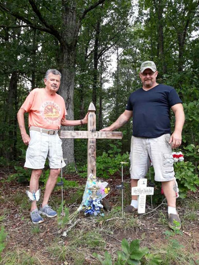Two men standing near a wooden cross memorial in a forest, remembering a late son and his heartbeat teddy bear.