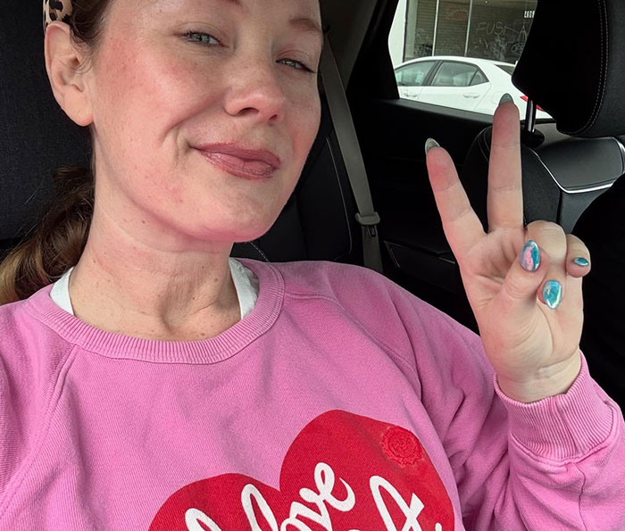 Woman in a pink sweatshirt making a peace sign inside a car, related to Tom Hanks breaking silence after daughter&rsquo;s claims.