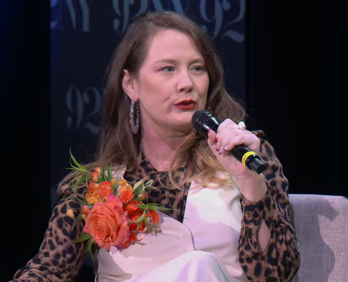 Woman holding microphone and orange flowers, speaking during a discussion about Tom Hanks and childhood struggles revealed by daughter.